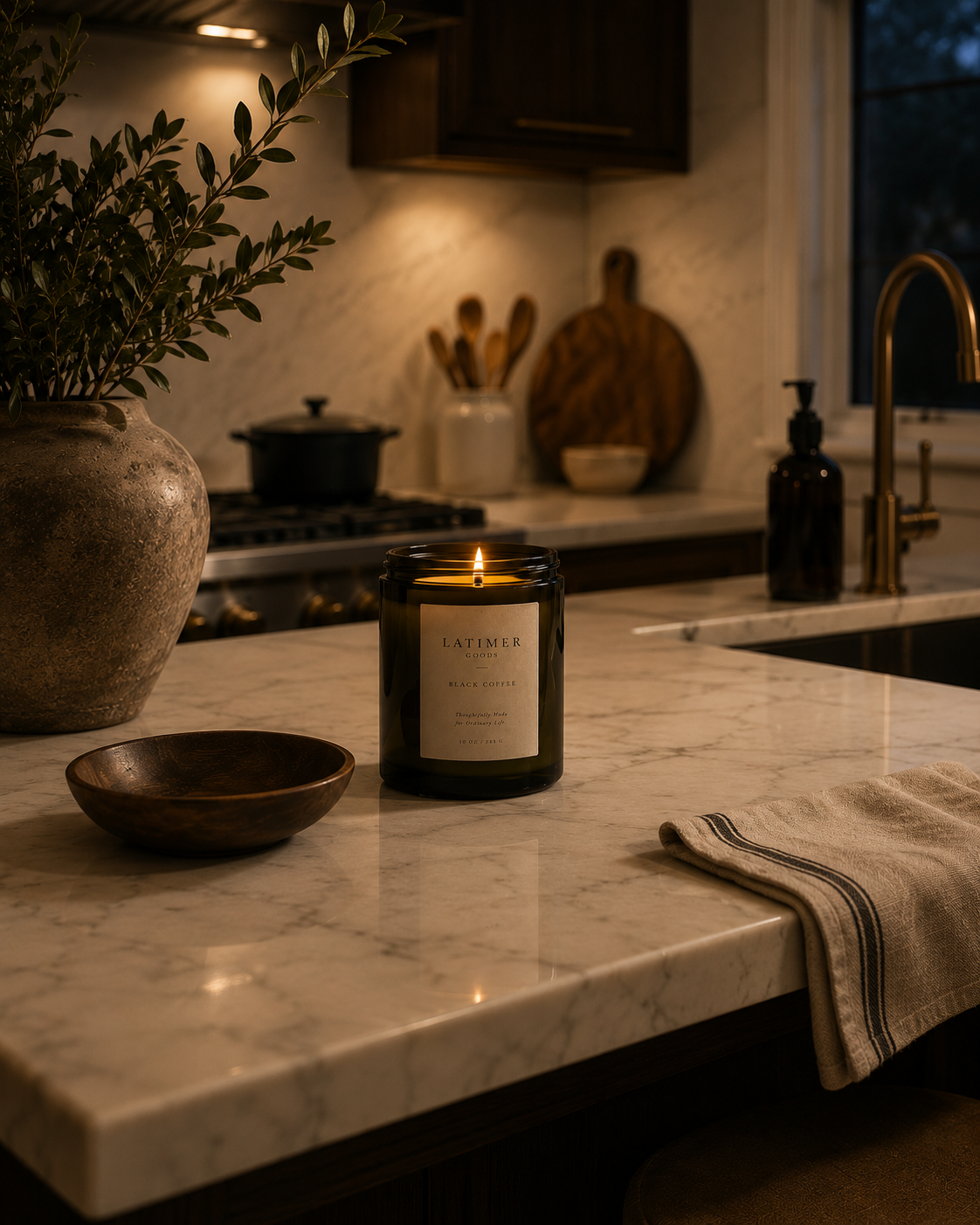 A lit candle in a dark glass jar sits on a marble kitchen island, surrounded by a wooden bowl, a folded towel, and a vase with greenery in a warmly lit kitchen setting.